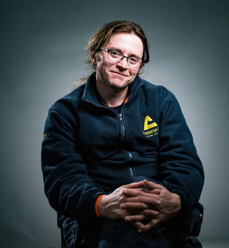 This is an image of Kat, a trans woman and HS2 worker, sitting in a studio. She is wearing a dark blue zip up jacket. Her hair is in a ponytail and she is wearing glasses. Her hands are folded on her knees and she is smiling.