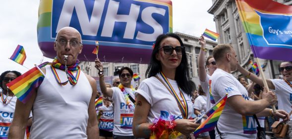 Protestors wearing rainbow accessories stand below an NHS Pride balloon.