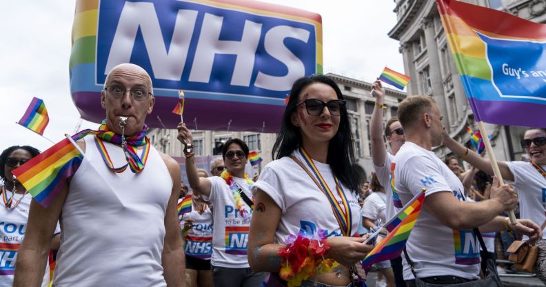 Protestors wearing rainbow accessories stand below an NHS Pride balloon.