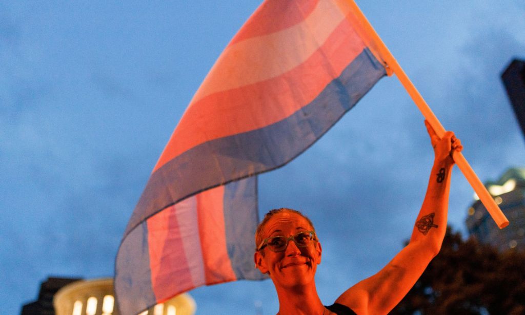 A person holds up a trans flag, both of which are bathed in an orange glow.