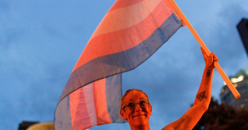 A person holds up a trans flag, both of which are bathed in an orange glow.
