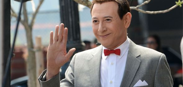 Pee-wee Herman actor Paul Reubens wearing a grey suit, white shirt and red bow tie. He is smiling and waving off camera.