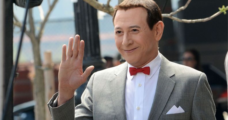 Pee-wee Herman actor Paul Reubens wearing a grey suit, white shirt and red bow tie. He is smiling and waving off camera.