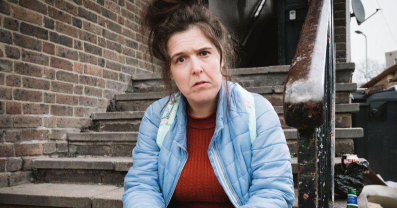 Comedian Rosie Jones as Emily in new sitcom Disability Benefits. She is staring with a questioning glance at the camera, while sitting on some steps in a back alley.