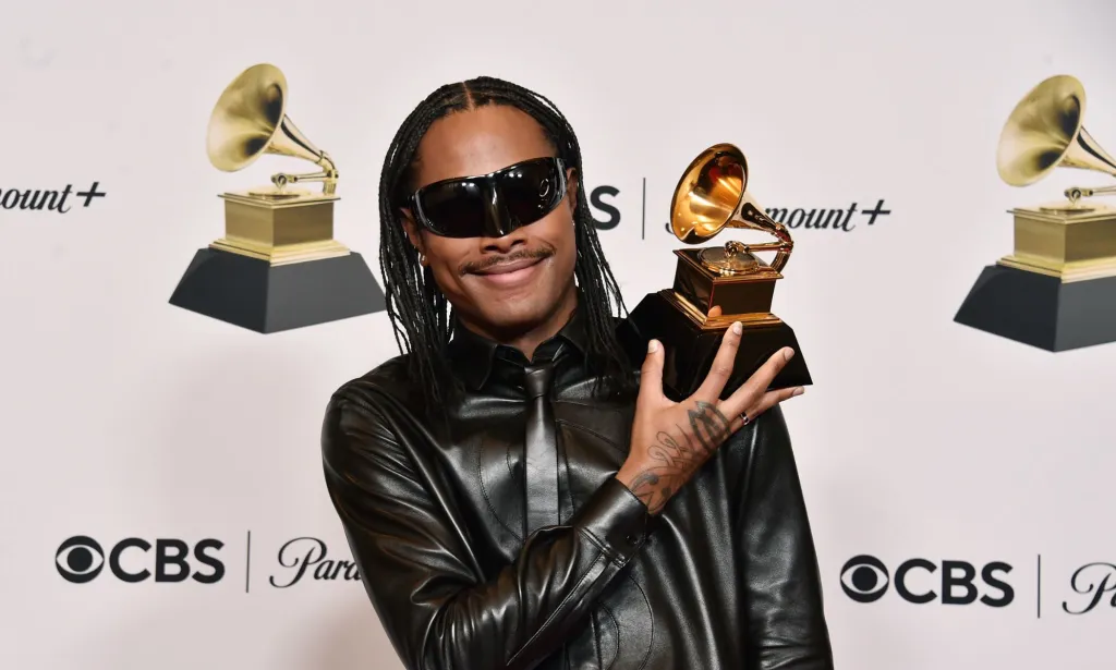 Steve Lacy wears a leather jacket and black sunglasses while smiling and holding his Grammy award.