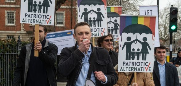 A man holds a megaphone while people behind him hold up anti-LGBTQ+ Patriotic Alternative signs.