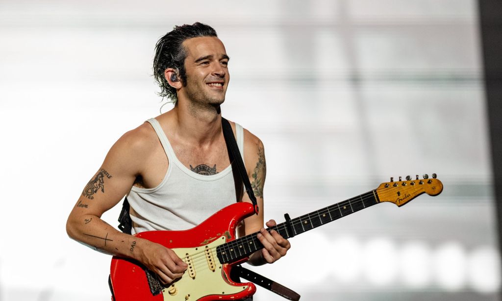 Matty Healy performs on stage in a white vest. He is playing a red electric guitar.