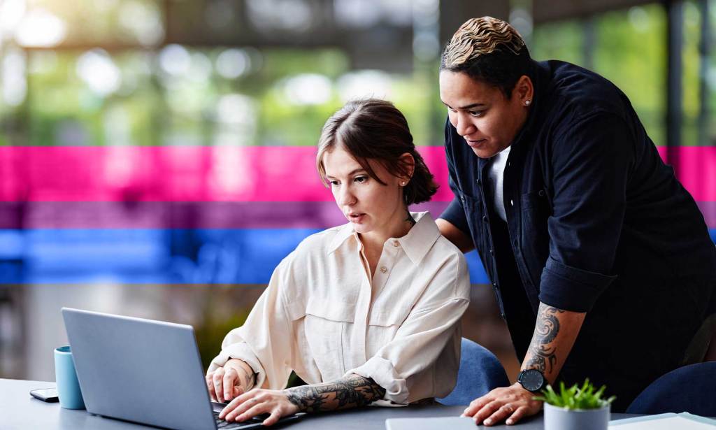 This is an image of two female presenting people at work. One person is sitting at a desk looking at a computer and they are looking at a computer. The other person is standing over them.