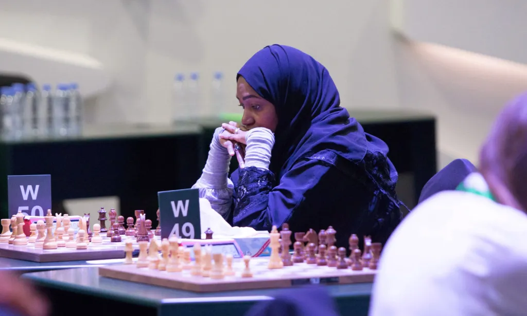 A woman wearing a blue hijab poses with her hands in front of her face as she thinks during a chess match