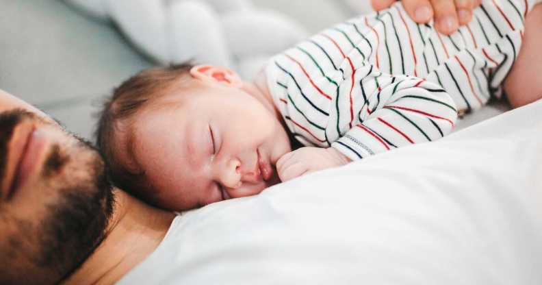 In this image, a man lies on a bed wearing a white t-shirt. His baby lies sound asleep on his chest as he cradles the child.