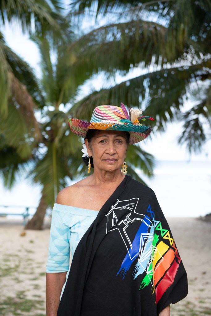 Pictured is Lady Tuaine Marsters is the spouse of The King's Representative to Cook Islands, Sir Thomas Marsters, and a long time, vocal supporter of LGBTI+ rights in the Cook Islands. She is standing on a beach wearing a hat and a shawl with a tree in the background.