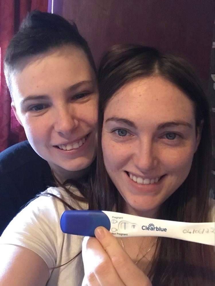 Queer couple Danielle and Natalie hold each other close as they hold up a pregnancy test to illustrate their fertility journey