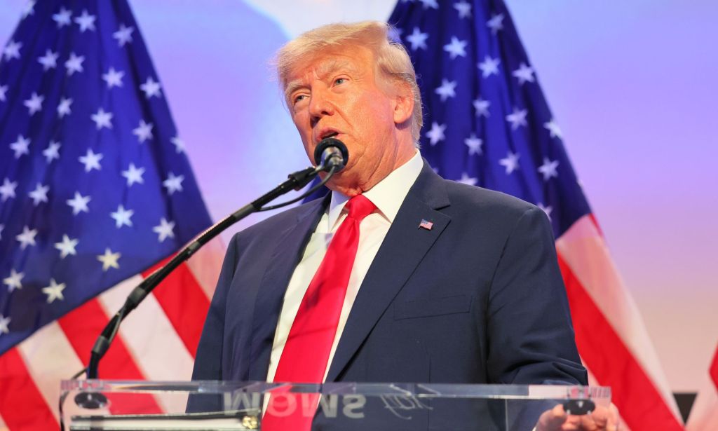 Former Republican president Donald Trump wears a white shirt, red tie and blue shirt as he speaks into a microphone at an event. Trump's presidential campaign has been marked by anti-trans talking points