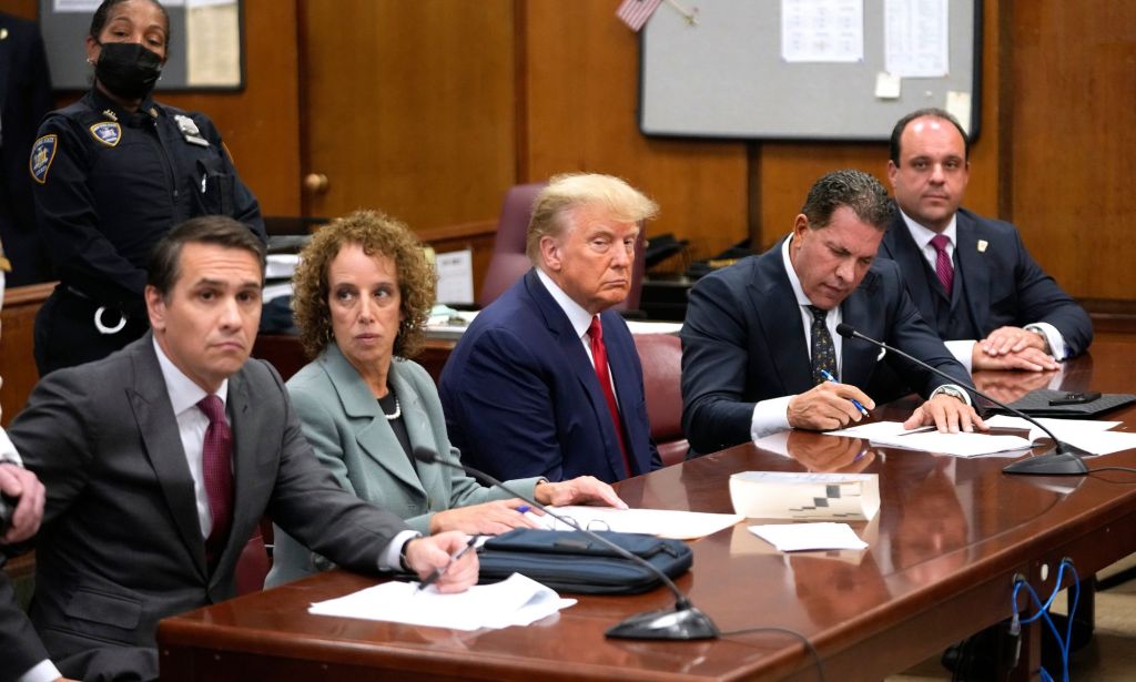 Former president Donald Trump wears a suit and tie while sitting at a table and surrounded by his legal team during an indictment hearing
