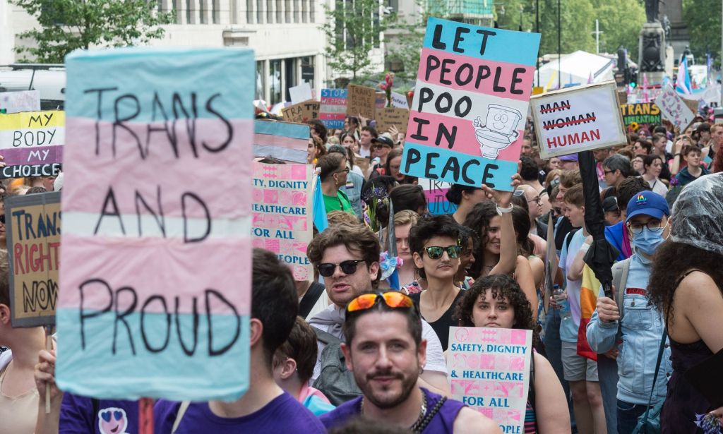 A person holds up a sign in the colours (pink, blue and white) of the trans Pride flag that reads 'Trans and proud' while another reads 'Let trans people poo in peace' specifically referencing efforts by the UK government to implement a trans bathroom ban/crackdown on gender-neutral toilets