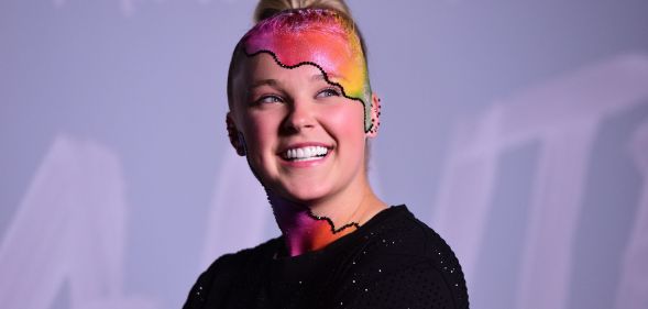 JoJo Siwa wears a black top as she poses with rainbow makeup on her head and neck