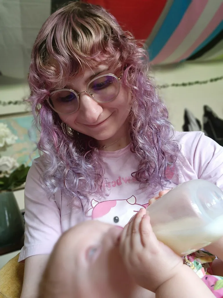 Naomi is pictured here feeding her child with pumped milk. She has pink hair and is wearing a pink t-shirt and stares down lovingly at her child.