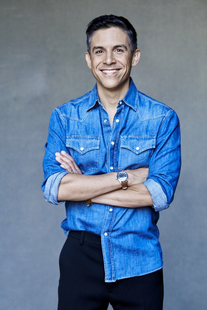 Matthew López in a blue shirt and dark jeans. He is stood against a grey background, smiling, with his arms folded.
