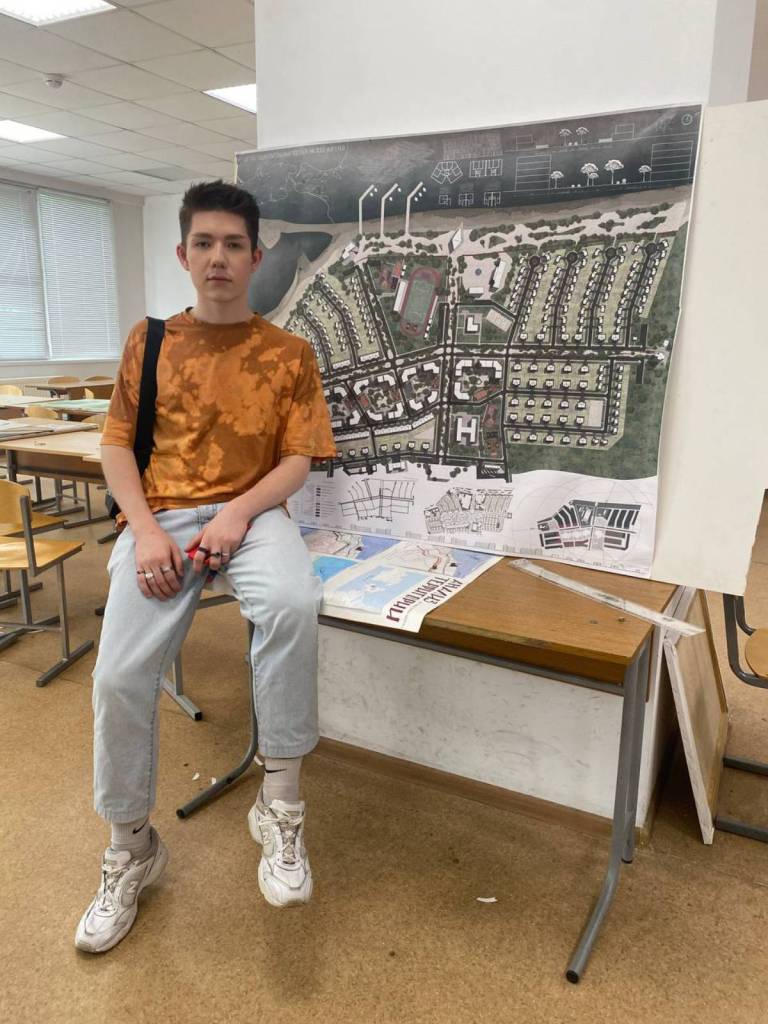 gay Russian university student, Max, sits on a table as he poses in front of a picture of a city