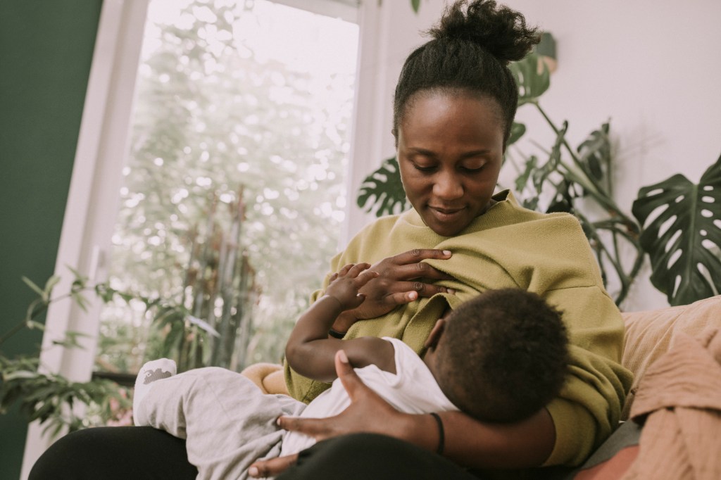 This stock image shows a Black woman breastfeeding her baby. She is wearing a green sweater which she has pulled up to feed her child.