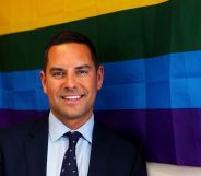 Openly gay Australian MP Alex Greenwich, who has received homophobic hate online, wears a suit and tie as he stands in front of a rainbow LGBTQ+ Pride flag
