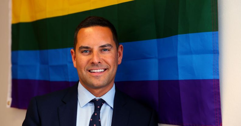 Openly gay Australian MP Alex Greenwich, who has received homophobic hate online, wears a suit and tie as he stands in front of a rainbow LGBTQ+ Pride flag