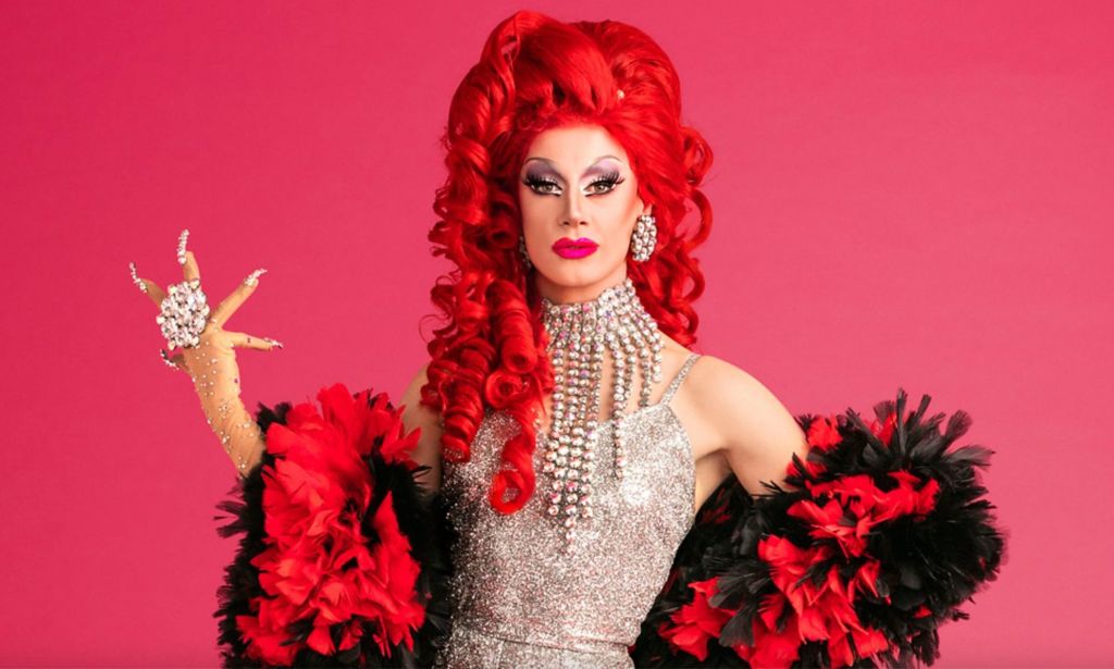 A promotional photo of Divina De Campo in Drag Race UK. She is wearing a red wig and a silver dress.