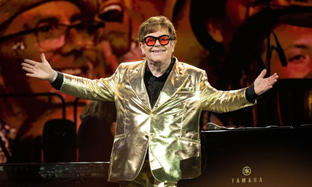 Elton John stands on stage wearing a gold suit