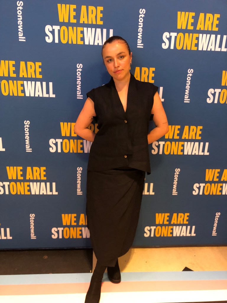 This is an image of a woman stand on a blue backdrop with the words "We are Stonewall" in yellow and white. She is wearing a black dress.