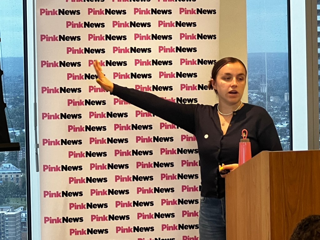 This is an image of a woman speaking. She is wearing a black sweater and is in front of a banner that reads PinkNews in pink and black colours.