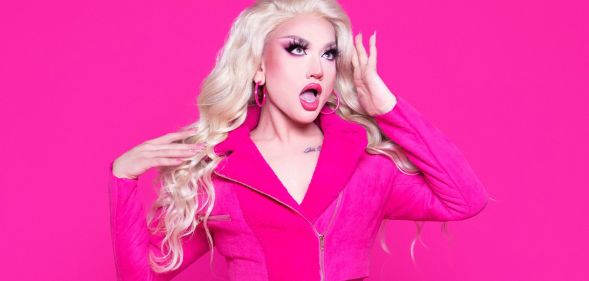 Drag star Lagoona Bloo in a pink jacket looking surprised holding her hand to her forehead. She is stood against a hot pink background.