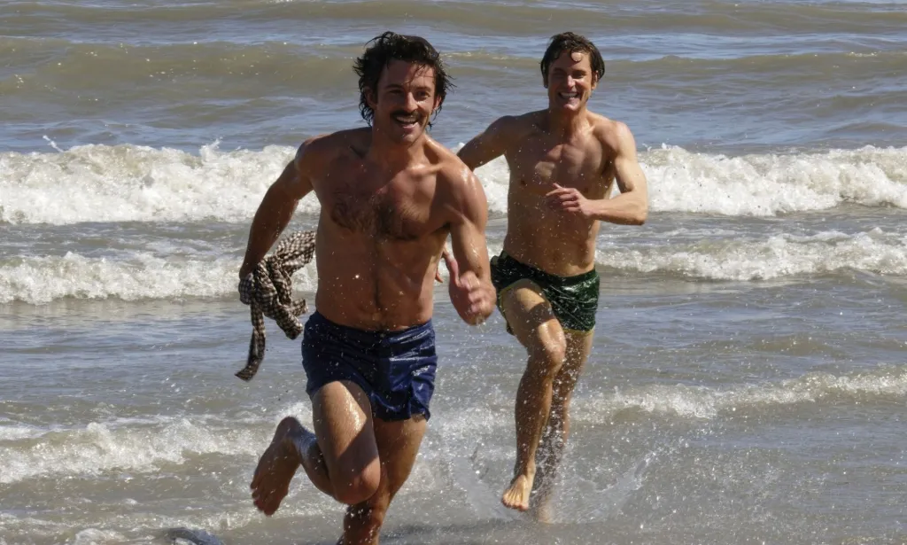 Jonathan Bailey and Matt Bailey chase each other on a beach in a still from Fellow Travelers.