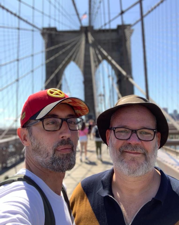 This is an image of two men on the Brooklyn Bridge. The man on the left is wearing a read baseball cap and a white tshirt. He also has glasses and a short beard. The man on the right has on a sun hat and is also wearing glasses.