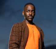 Ncuti Gatwa in a promotional photo for Doctor Who. He is wearing a brown and black chequered jacket and orange top with a necklace. He is looking off camera. The sky is behind him.