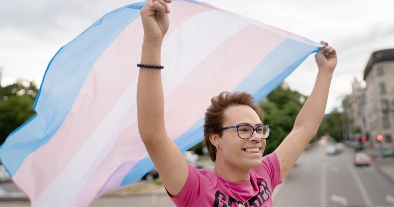 Person wearing a pink shirt and smiling, waving a transgender Pride flag behind them