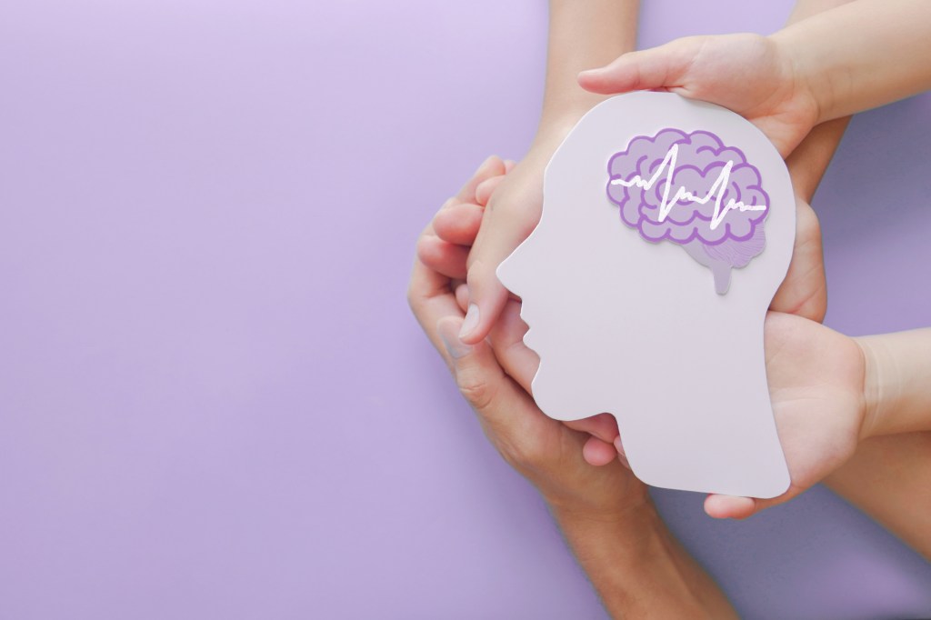 This picture shows an adult and a child's hands holding a cutout of a person's head with a purple brain. The image is set against a purple background.