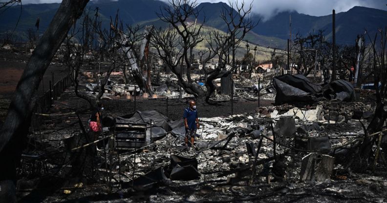 A ground view of the devastation left behind in Maui, Hawaii after wildfires burned down Lahaina. In the picture, there are two people shifting through the rubble, blackened trees and the remains of buildings