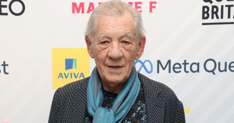 Ian McKellen wears a dark shirt, blue patterned silk scarf and dark grey jacket as he stares towards the camera