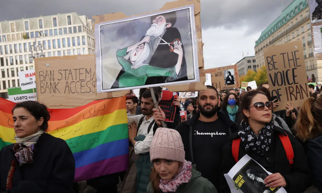 People gather together in protest one year after the death of Mahsa Amini, a Kurdish-Iranian woman who died in the custody of Iran's 'morality police'. One person holds up a rainbow LGBTQ+ pride flag while others hold up signs in protest against Iran's regime