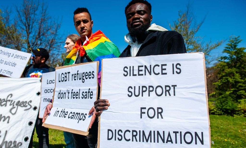 People hold up signs bringing awareness to the plight of LGBTQ+ people seeking asylum abroad including a sign reading 'silence is support for discrimination' and another reading 'LGBT refugees don't feel safe in the camps'
