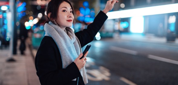 Woman arranging taxi service using mobile app on smart phone, standing on the city street, with illuminated street lights in the background.