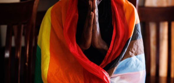 A person in Uganda wears an LGBTQ+ Progressive Pride flag draped around their shoulders as they hold their hands in front of them in prayer