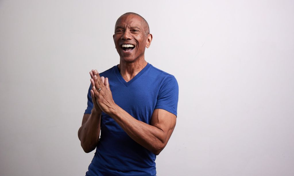 Larry Duplechan in a blue t-shirt laughing with his hands pressed together.