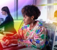 This is an image of a Black woman working at a computer. She is wearing a floral top. There is a creative overlay with the Pride colours over the top