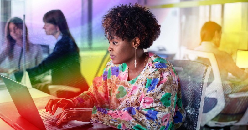 This is an image of a Black woman working at a computer. She is wearing a floral top. There is a creative overlay with the Pride colours over the top
