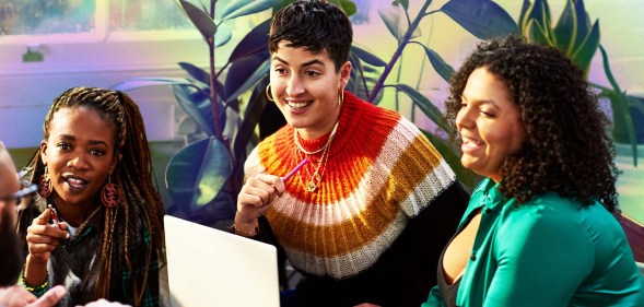 This is an image of a diverse goup of colleagues. On the left there is a Black woman with long braids and a black jumper. In the middle is a white woman with short hair sitting over a laptop. The woman on the right has dark hair and is wearing green.
