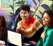 This is an image of a diverse goup of colleagues. On the left there is a Black woman with long braids and a black jumper. In the middle is a white woman with short hair sitting over a laptop. The woman on the right has dark hair and is wearing green.