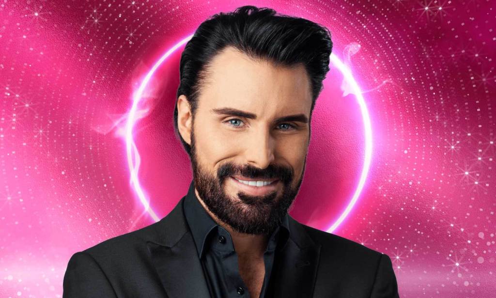 This is an image of Rylan Clark. He has dark hair, a short beard. He is wearing a black suit and is smiling.