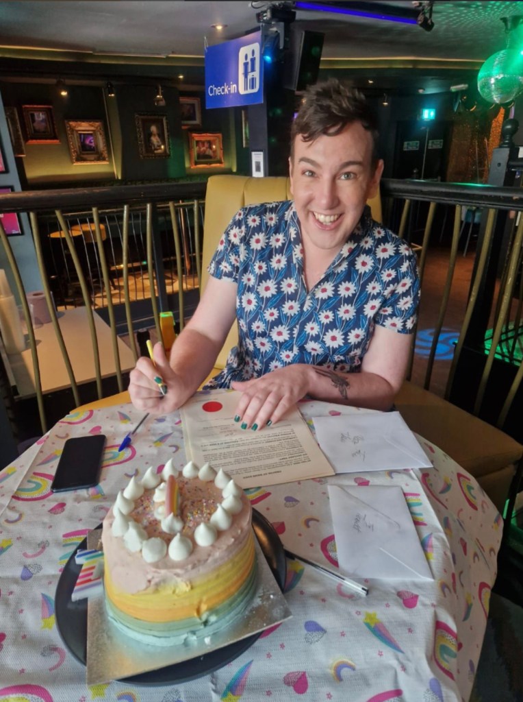 This is an image of a trans and non-binary person signing a document. There is a cake on the table. She is wearing a blue dress with white accents. 