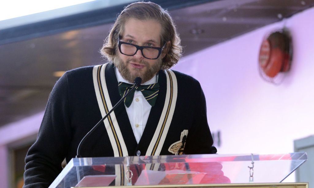Bryan Fuller wears a white shirt, black bow tie and striped jumper as he speaks into a microphone